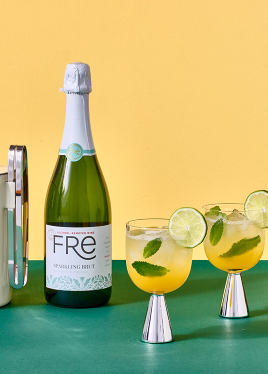 Bottle of FRE Alcohol-Removed sparkling brut white wine next to two glasses filled with yellow cocktails garnished with herb leaves and slices of lime