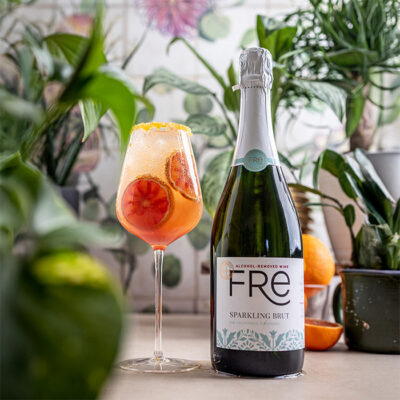 color image of a bottle of FRE Alcohol-Removed Sparkling Brut with an orange cocktail in front of green plants