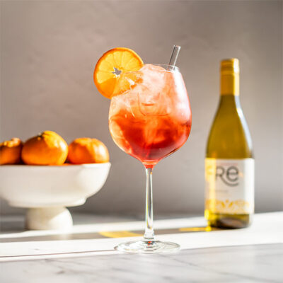 sunny image of a bright orange mocktail with a bottle of FRE Alcohol-Removed Chardonnay in the background