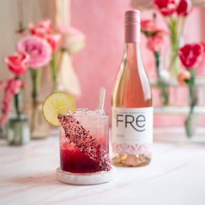 color image pink flowers in the background with a bottle of FRE Alcohol-Removed Rose wine with a hot pink cocktail with candy rim