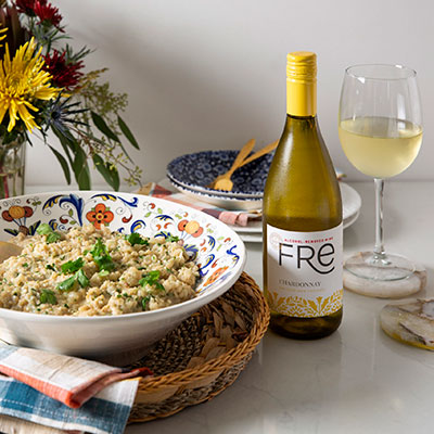 Large bowl of Creamy Coconut-Cilantro Cauli-Rice on a table with flowers in the background, wine glasses, and a bottle of FRE alcohol-removed Chardonnay white wine