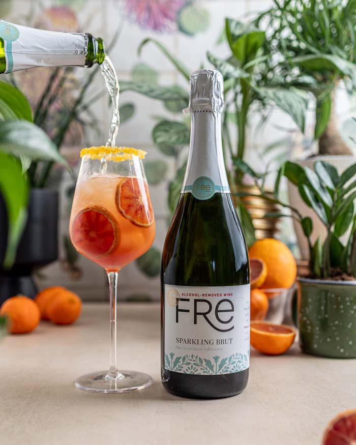 color image of a bottle of FRE Alcohol-Removed Sparkling Brut with an orange cocktail in front of green plants