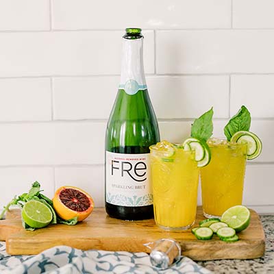 Color image of a bottle of FRE Alcohol-Removed Sparkling Brut with 2 yellow cocktails with a basil and cucumber garnish