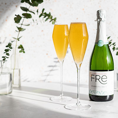 Two champagne flutes with orange-colored bellini mocktails in them, and a bottle of Fre Alcohol-Removed Sparkling brut non-alcoholic white wine