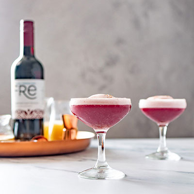 Two coupe cocktail glasses containing dusky pink apple sour mocktails with a bottle of Fre Alcohol-Removed Cabernet Sauvignon non-alcoholic red wine in the background