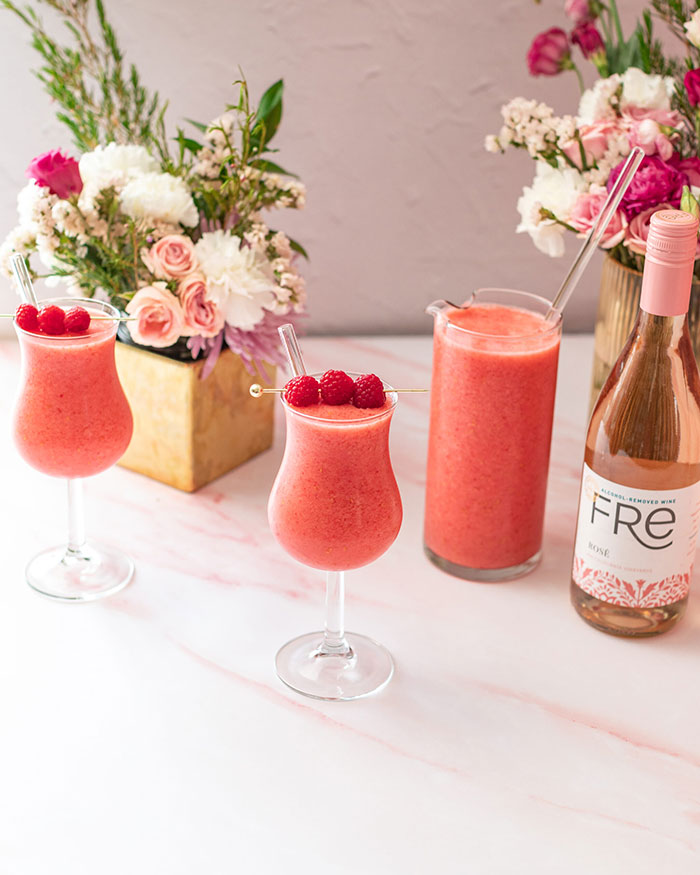 Two glasses of pink raspberry frosé mocktail garnished with raspberries, with a bottle of Fre non-alcoholic rosé wine