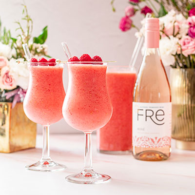 Two glasses of pink raspberry frosé mocktail garnished with raspberries, with a bottle of Fre non-alcoholic rosé wine in the background