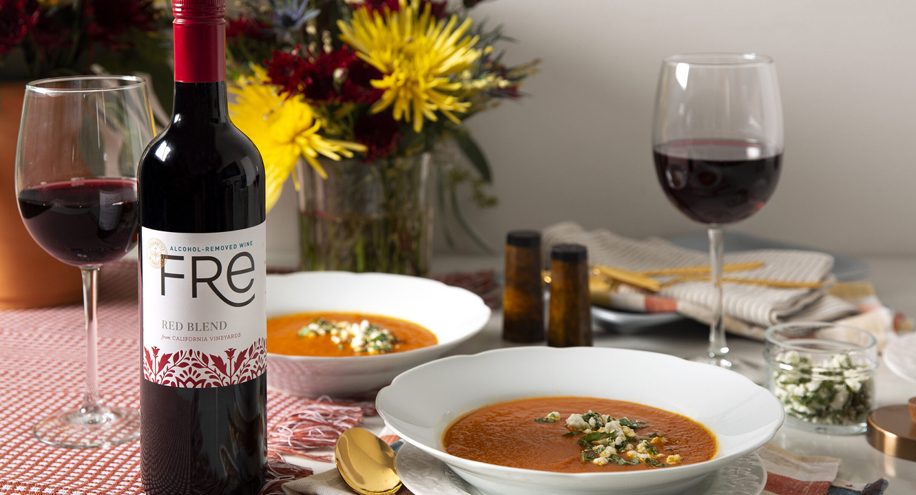 Two bowls of Turmeric Roasted Tomato Soup with Goat Cheese, alongside wine glasses and a bottle of FRE alcohol-removed Red wine