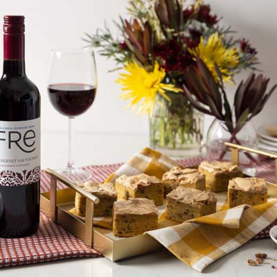 A gold tray of blond brownies on a cloth napkin with a bottle of FRE Alcohol-Removed Cabernet Sauvignon and a wine glass filled with red wine, with a vase of flowers in the background