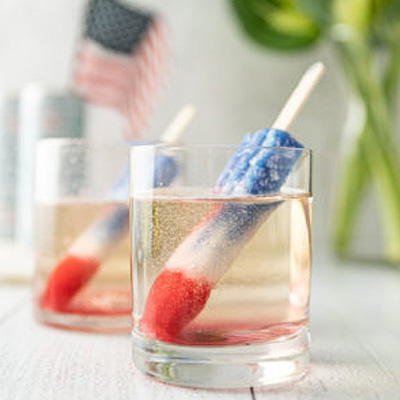 Two rock glasses on a wood surface that contain sparkling wine and frozen red white and blue pops, with a blurred United States flag and greenery in the background
