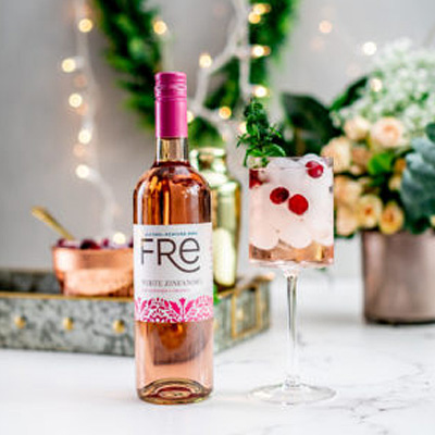 FRE Alcohol-Removed White Zinfandel bottle with a pink fancy desert cocktail next to it on a marble surface, with blurred flowers, a tray with accoutrements, string lights, and greenery in the background
