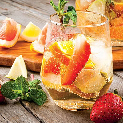 Close up of a cocktail with clear liquid and pieces of fresh fruit on a wood table, with a wood cutting board in the background and fresh cut citrus fruit, mint leaves, and a strawberry in the foreground