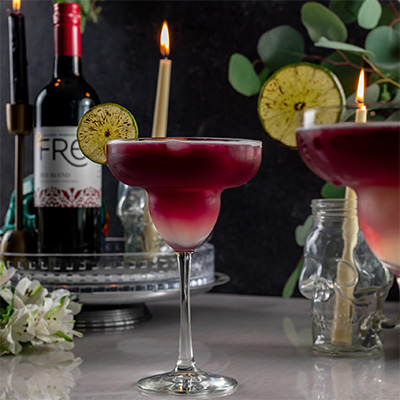 Fancy cocktail glasses filled with red colored cocktail with candles, greenery, glassware, and FRE Alcohol-Removed Red Blend bottle in the background