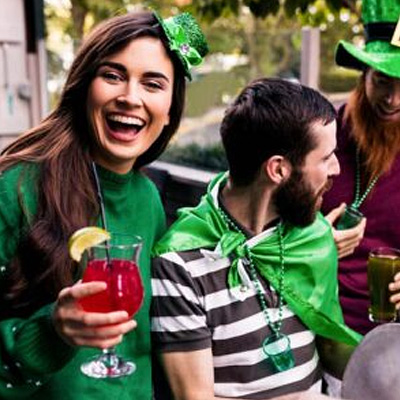 A smiling woman dressed in a green jumper and small sparkly green hat holds a mocktail, next to a man wearing a green cape, with another man dressed as a leprechaun in the background