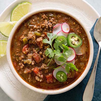 Bird's eye view of a bowl of chili with cilantro, sliced radish, and sliced jalapeno toppings. with sliced limes garnish