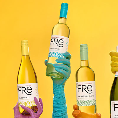 Hands wearing long evening wear-type gloves in bright colors hold various bottles of white Fre non-alcoholic wines in front of a solid yellow background