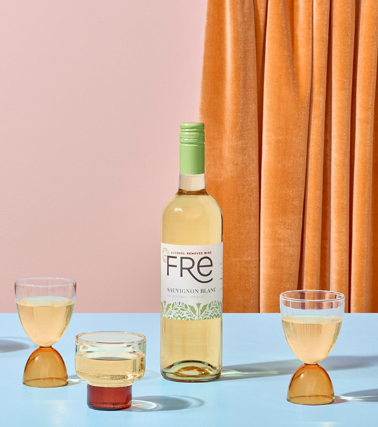 A bottle of Fre non-alcoholic sauvignon blanc white wine, with three retro wine glasses with colorful wide stems and an orange curtain in the background to give a stylish retro sixties effect
