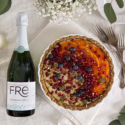 Bird's eye view of a colorful tart with blueberries, pomegranate, and other toppings, with a bottle of FRE Alcohol-Removed Sparkling Brut and some Baby's Breath flowers