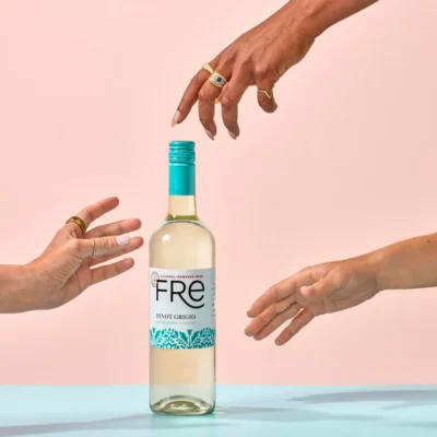 Bottle of FRE Alcohol-Removed Pinot Grigio on light blue surface and peach colored background with three hands coming in from the sides to grasp the bottle