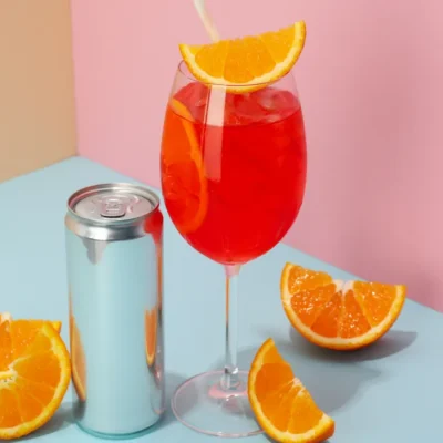 A brightly colored orange red cocktail inside a wine glass on a light blue surface and a silver can next to it, with fresh orange slices, and a tan and pink background