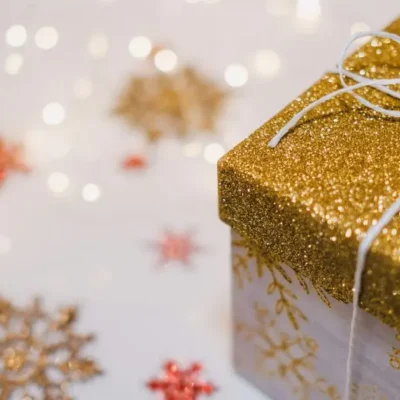 A wrapped gift with gold glitter wrapping and white twine bow with a blurred light festive background with sparkles and stars