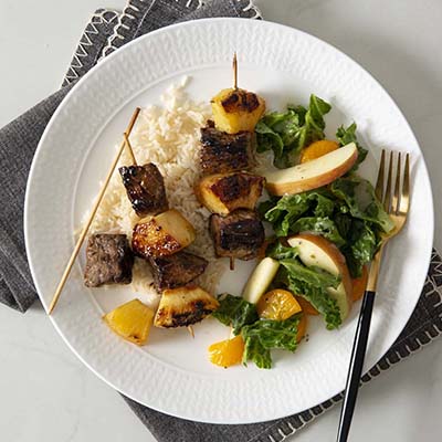 Bird's eye view of a plate of rice with steak and pineapple kabobs and a side salad with greens and sliced apples