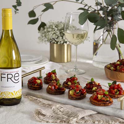 A bottle of FRE Alcohol-Removed Chardonnay to the left with appetizers on a marble tray and a wine glass filled with white wine, with flowers in a vase and greenery in the background